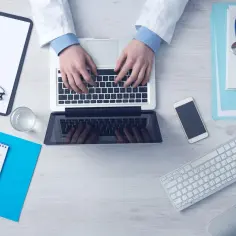 Lekarz wpisuje dane pacjenta na laptopie. Obok leżą stetoskop, telefon, notatnik z termometrem. Ważne jest skierowanie na badania.