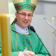 Uśmiechnięty prymas polski w zielonej szacie i mitrze, przed mikrofonem, obok świec i krucyfiksu.