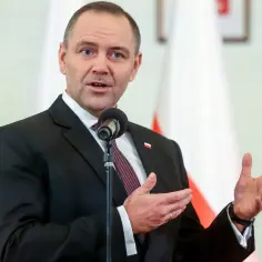 Premier Polski w garniturze, przemawiający przy mikrofonie na tle flag.