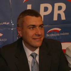 Mężczyzna w garniturze, z niebieskim krawatem, na tle banera z napisem "LPR" i hasłami "Mądra edukacja" oraz "Bezpieczna rodzina".