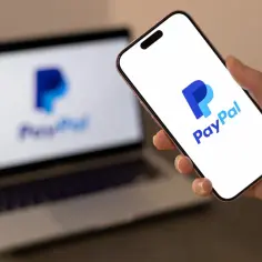Dłoń trzyma smartfon z logo PayPal, a w tle widać laptop z tym samym logo. Czy PayPal jest bezpieczny? Tak, to popularna i bezpieczna platforma płatności online.