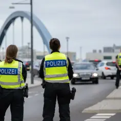 Funkcjonariusze policji w kamizelkach odblaskowych "POLIZEI" prowadzą kontrole na granicy niemieckiej. W tle widać most i ruch samochodowy.