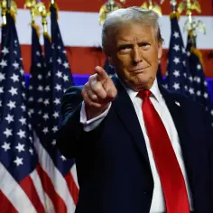 Donald Trump, prezydent USA, wskazuje palcem na widza, stojąc przed amerykańskimi flagami.