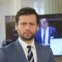 Minister sportu w niebieskiej marynarce i krawacie w paski, z zarostem, patrzy prosto w obiektyw.