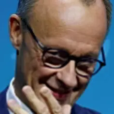 Zbliżenie na twarz mężczyzny w okularach, uśmiechającego się i dotykającego ust dłonią.