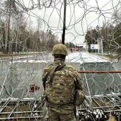 Żołnierz w mundurze obserwuje teren przez zasieki z drutu kolczastego. Widoczny jest znak graniczny, sugerujący, że to granica polsko-białoruska.