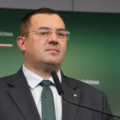 Minister rolnictwa w garniturze i krawacie przemawia przy mikrofonie na tle zielonej ściany z logo ministerstwa.