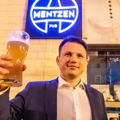 Mężczyzna w garniturze wznosi kufel piwa przed neonem "Mentzen Pub".