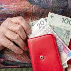 Dłoń starszej osoby wkłada banknoty 20 i 100 zł do czerwonego portfela. Czerwcowe emerytury wreszcie w rękach.