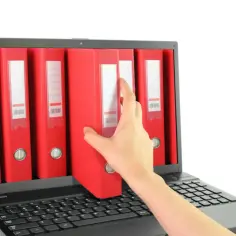 Dłoń sięga po czerwoną teczkę z cyfrowego archiwum na ekranie laptopa. To symbol cyfryzacji dokumentacji, np. w ramach zfśs.