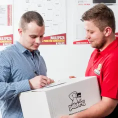 Kurier w czerwonej koszulce przekazuje paczkę, a klient podpisuje dokument. Zastanawiasz się, ile zarabia kurier?