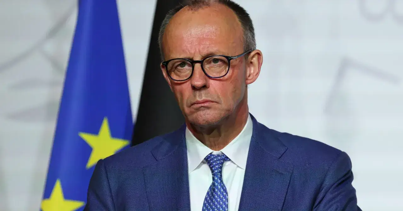 Kanclerz Niemiec w okularach, w garniturze i krawacie z flamingami, na tle flagi UE.