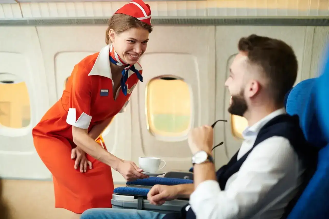 Uśmiechnięta stewardessa w czerwonym stroju podaje kawę pasażerowi w samolocie.
