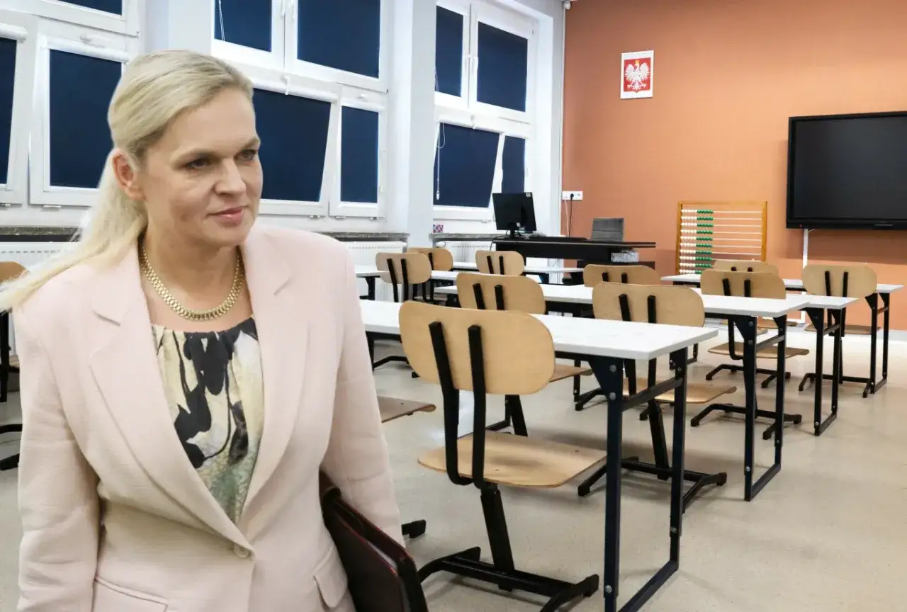 Minister edukacji w różowym żakiecie stoi w pustej sali lekcyjnej.