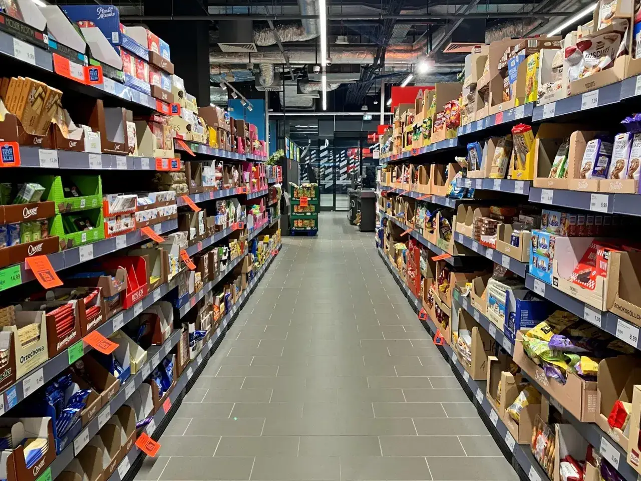 Alejka sklepowa pełna produktów FMCG, od słodyczy po przekąski. Półki uginają się pod ciężarem różnorodnych towarów, tworząc labirynt dla kupujących.