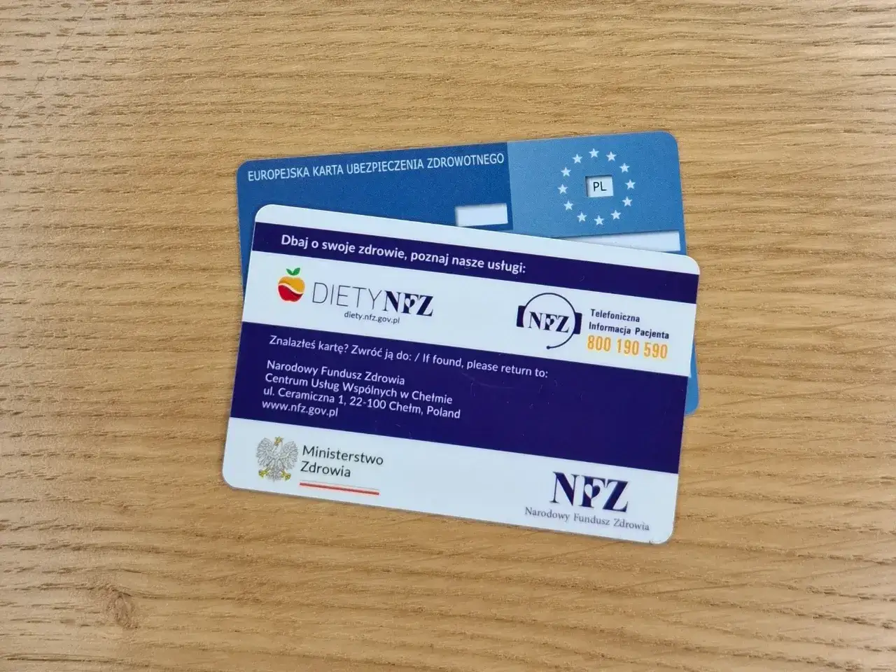 Karta EKUZ i karta Diety NFZ na drewnianym tle. Dwie karty ubezpieczenia zdrowotnego, jedna z symbolem Unii Europejskiej, druga z informacjami o diecie i kontaktem do NFZ.