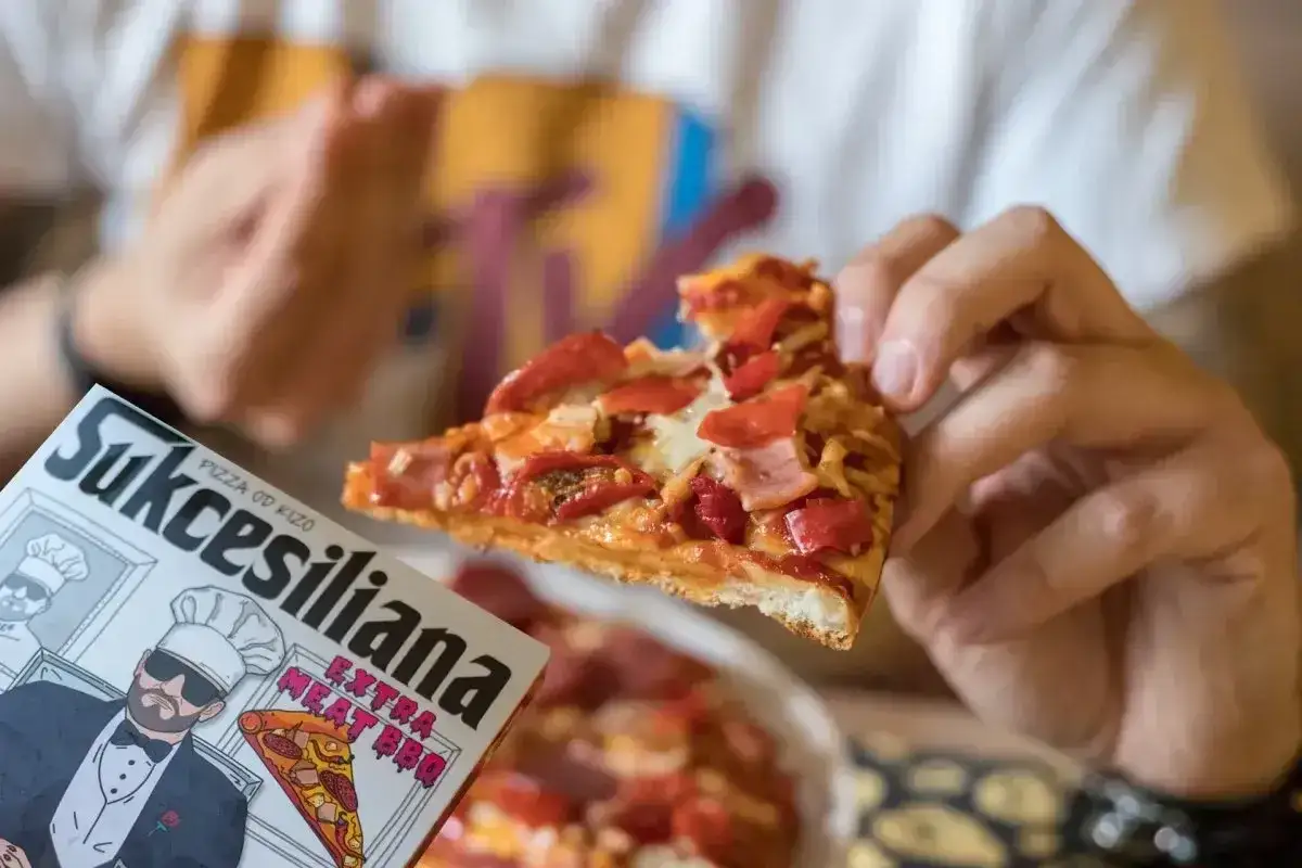 Ręce trzymają kawałek pysznej pizzy od Kizo. Obok pudełko z napisem 